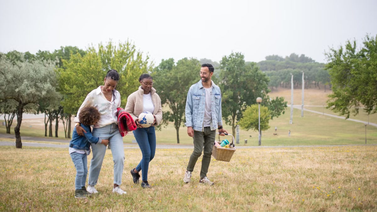 A joyful family walking through a park on a sunny day, carrying a picnic basket and a ball.